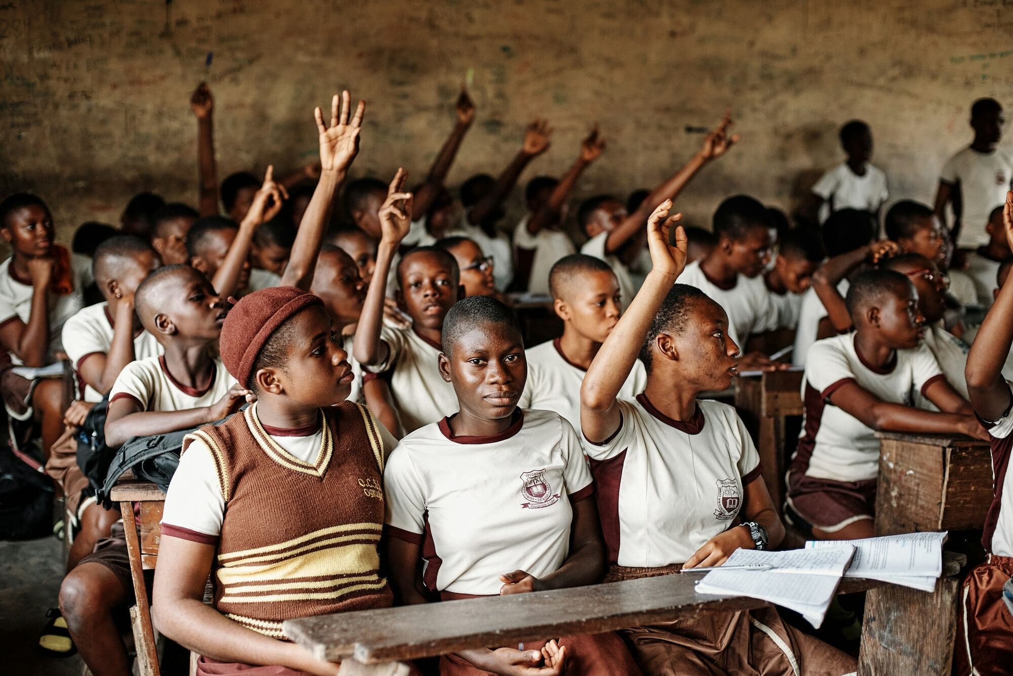 Secondary school students in cream and maroon uniforms raise their hands in a busy classroom