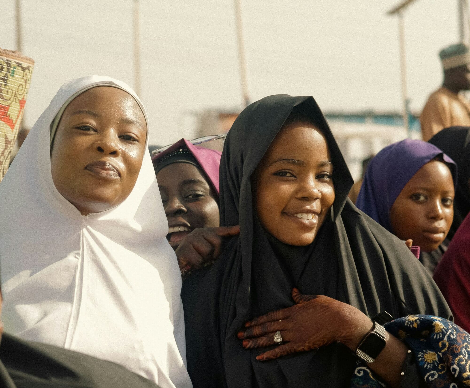 Smiling women and girls in white, black and purple headscarves stand close together at a sunny outdoor gathering