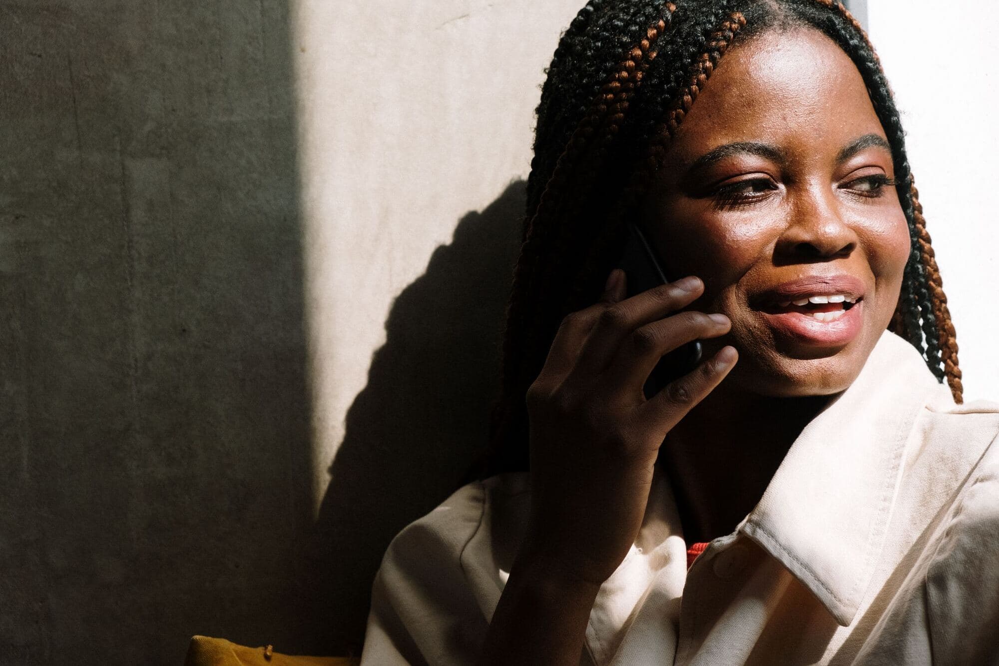 A smiling young woman with braids talks on a mobile phone in warm sunlight beside a wall