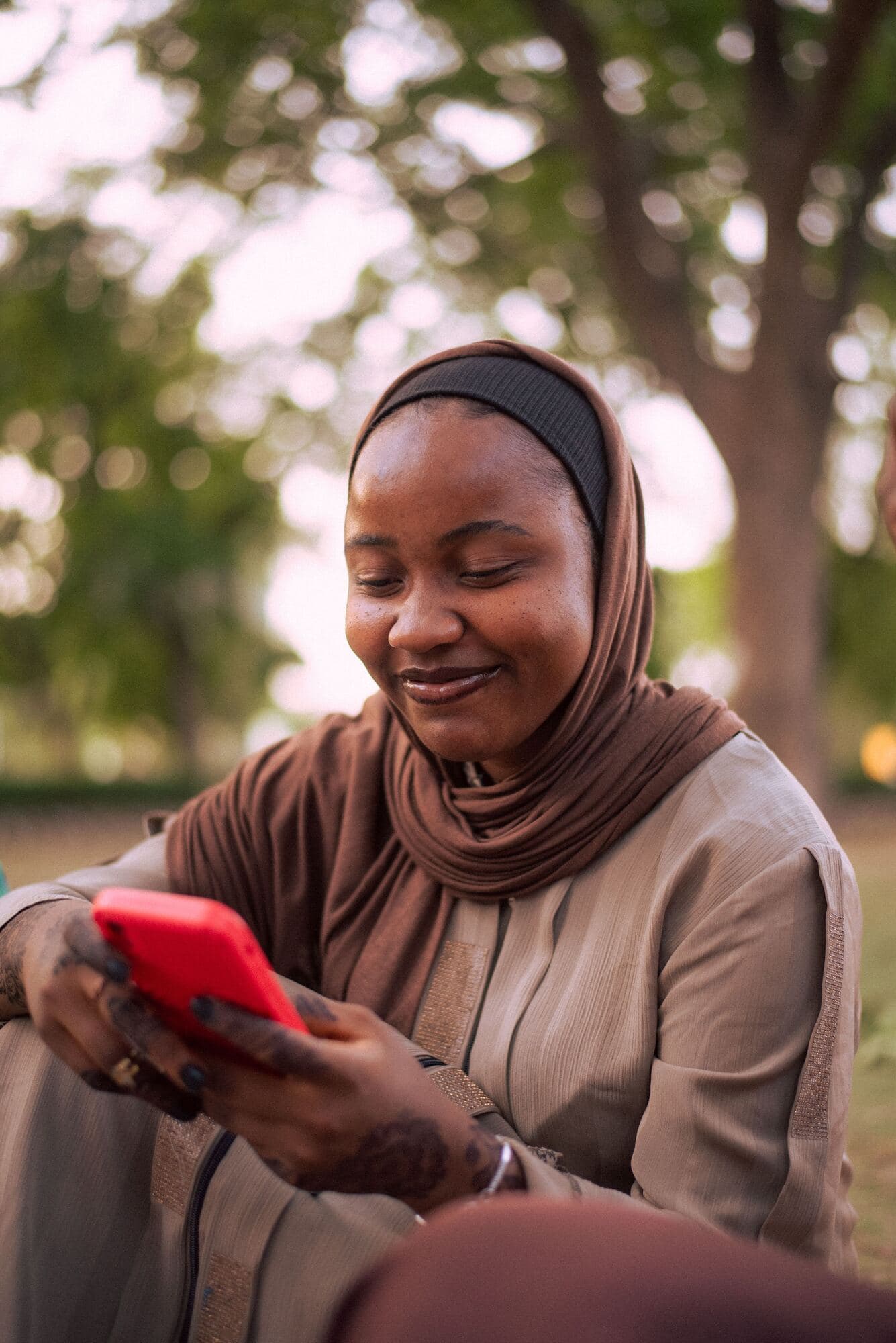 A smiling young woman in a brown headscarf looks at a red phone while sitting outdoors under trees