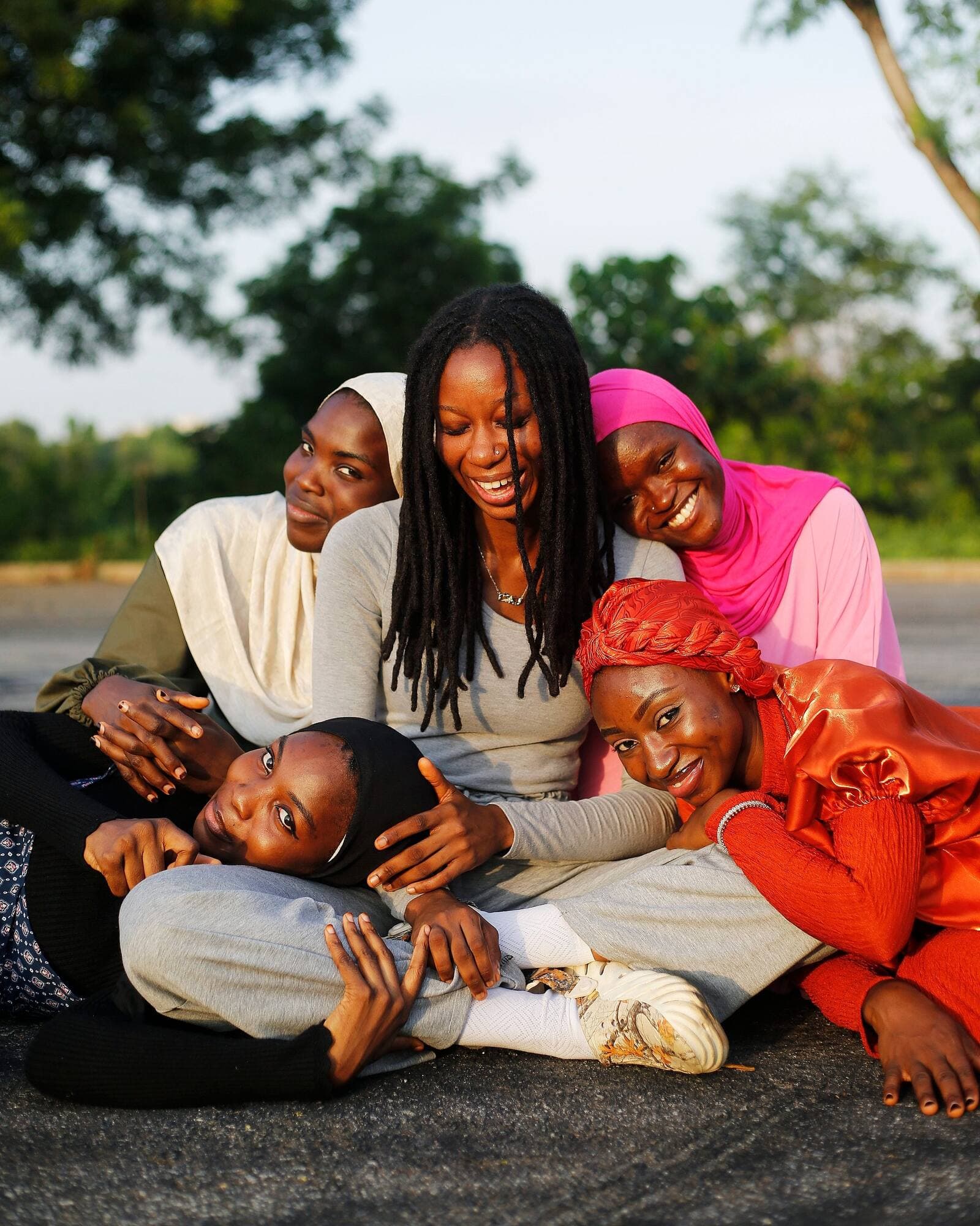 Five young women, some in headscarves, laugh and lean on each other outdoors in warm golden sunlight