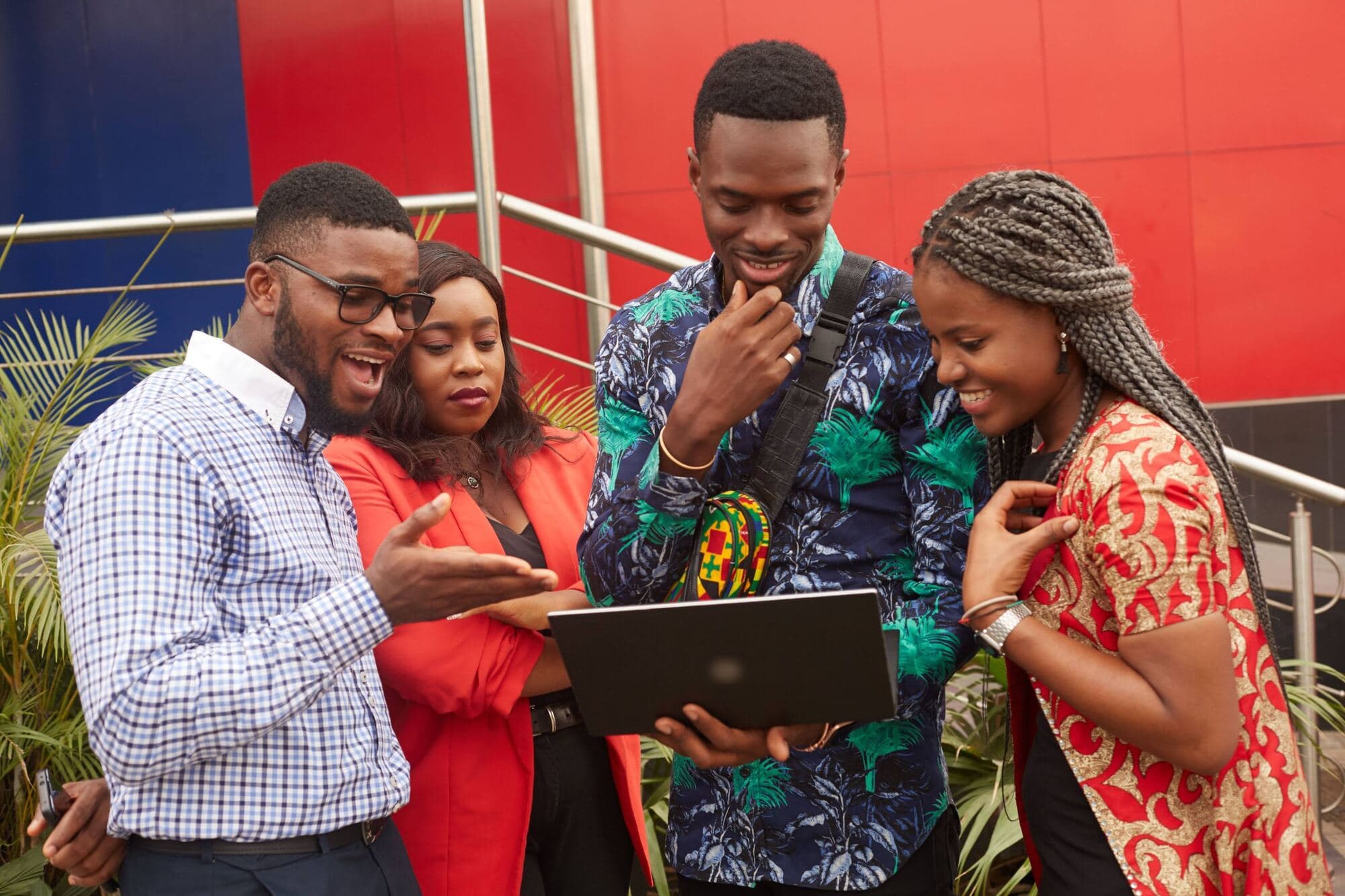 Four young professionals smile as they look at a laptop together outdoors against a red wall