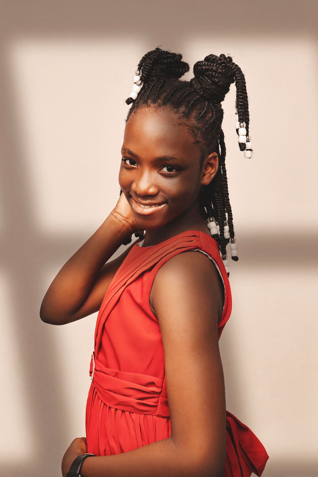A smiling girl in a red dress with beaded braids poses confidently in warm light