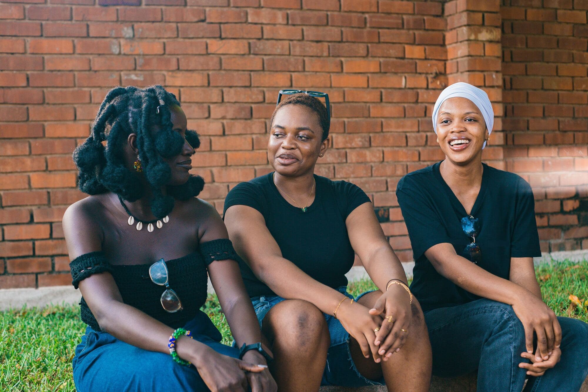 Three young women in black tops sit on a step by a red brick wall, chatting and laughing